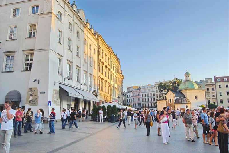 28 - A lively view of the Main Market Square in Kraków, featuring pedestrians strolling past historic European architecture and the distinctive cupola of the Church of St. Wojciech